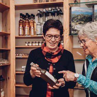 Two women are looking at a bottle together in a store. Shelves with additional products can be seen in the background. | © Carolin Ries | www.calejofotografie.de