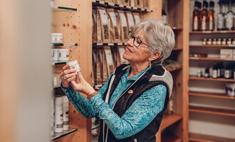 An elderly woman is looking at a product in a store. The room is well-lit and full of natural products. | © Carolin Ries | www.calejofotografie.de
