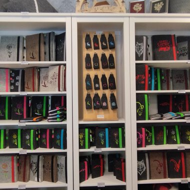 Eine Regalwand mit vielen Notizbüchern in verschiedenen Farben und Designs. Darunter befinden sich farbige Stifte und weitere Schreibwaren. | © Chrömle Schönes aus Filz | N. Lughammer
