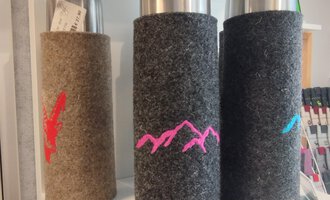 Drei Thermosflaschen mit Filzüberzügen in verschiedenen Farben stehen auf einem Regal. Die Flaschen haben farbige Bergmotive auf den Überzügen. | © Chrömle Schönes aus Filz | N. Lughammer