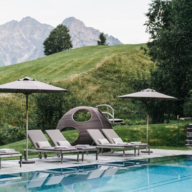 A pool area with loungers and sun umbrellas. In the background, gentle hills and mountains can be seen.
