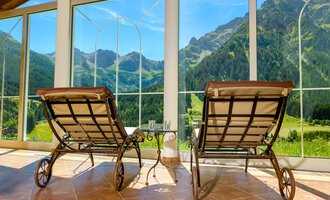 Zwei Liegestühle stehen vor großen Fenstern mit Blick auf grüne Berge. Der Raum wirkt einladend und hell. | © Hotel Leitner