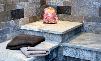 Ein modernes Saunainterior mit grauen Steinwänden. Auf einem der Steine liegt ein rosa Salzblock und einige Frotteehandtücher. | © Hotel Leitner