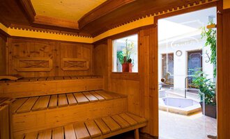 Eine gemütliche Sauna aus Holz mit einer Bank und großen Fenstern. Durch die Tür sieht man einen kleinen Innenpool und Pflanzen im Hintergrund. | © Hotel Leitner