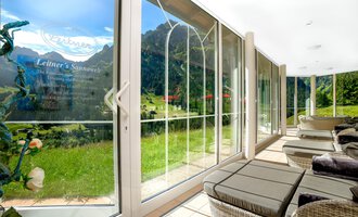 Ein moderner Raum mit großen Fenstern, die einen Blick auf eine grüne Landschaft und Berge bieten. Gemütliche Liegen laden zum Entspannen ein. | © Hotel Leitner