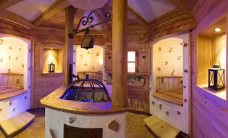 A cozy wellness area with wooden paneling and soft lighting. In the center, there is a round fountain surrounded by seating. | © Hotel Leitner