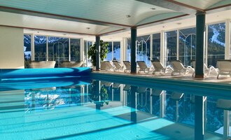 A bright indoor pool with clear water and loungers beside it. Large windows let in plenty of daylight and offer a view outside. | © Hotel Leitner