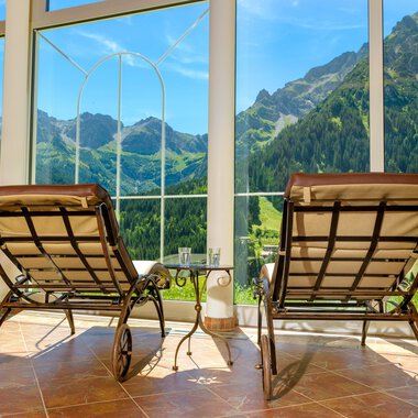 Two lounge chairs are positioned in front of large windows overlooking green mountains. The room feels inviting and bright. | © Hotel Leitner