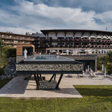 Ein modernes Hotel mit einem großen, gläsernen Pool steht im Vordergrund. Die umliegende Landschaft ist grün und einladend. | © A-ROSA Ifen Hotel Kleinwalsertal