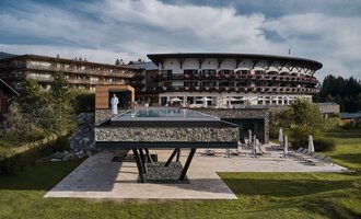 Ein modernes Hotel mit einem großen, gläsernen Pool steht im Vordergrund. Die umliegende Landschaft ist grün und einladend. | © A-ROSA Ifen Hotel Kleinwalsertal