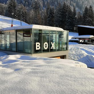 Ein modernes, gläsernes Gebäude mit der Aufschrift "BOX" steht inmitten einer schneebedeckten Landschaft. Im Hintergrund sind schneebedeckte Bäume und weitere Gebäude sichtbar. | © Sport Kessler GmbH