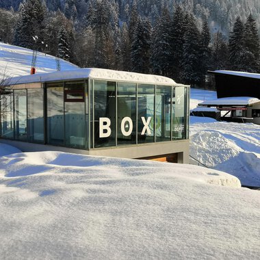 A modern glass building with the inscription "BOX" stands amidst a snow-covered landscape. In the background, snow-covered trees and additional buildings can be seen. | © Sport Kessler GmbH