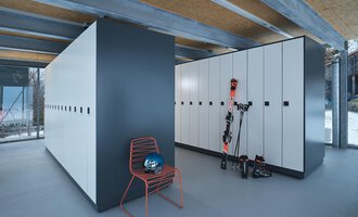 Ein moderner Skiraum mit weißen Schließfächern und einem roten Stuhl. An der Wand stehen Skiausrüstung und ein Helm. | © Sport Kessler GmbH
