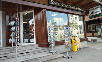 Ein Geschäft namens „Der bunte Laden“ mit großen Fenstern und einem einladenden Eingang. Draußen stehen mehrere Ständer mit Postkarten und Souvenirs. | © Der Bunte Laden | Romana Volderauer