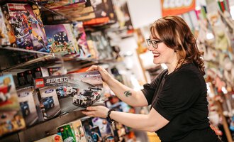 A woman is holding a LEGO Speed series set in a toy store. She smiles as she looks at the shelves with various LEGO sets. | © Sascha Gast, Dein Fotograf