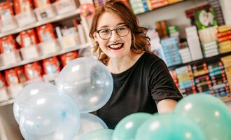 A young woman with glasses smiling while holding colorful balloons. In the background, shelves with books are visible. | © Sascha Gast