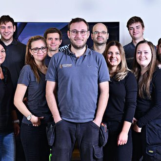 Eine Gruppe von zwölf Personen steht zusammen und lächelt. Sie tragen ähnliche graue Arbeitskleidung und befinden sich in einem hellen Raum. | © Der Elektriker Markus Leitgeb GmbH