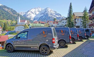 Vier Lieferwagen stehen auf einem gepflasterten Platz mit einer malerischen Bergkulisse. Im Hintergrund sind grüne Wiesen, Häuser und eine Kirche sichtbar. | © Der Elektriker Markus Leitgeb GmbH