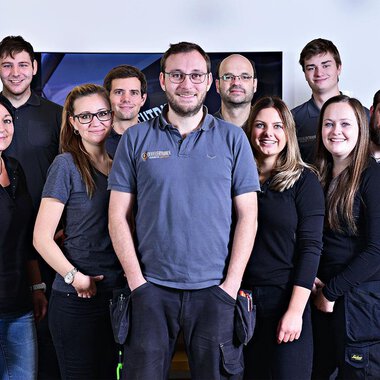 A group of twelve people stands together and smiles. They are wearing similar gray work clothing and are in a bright room. | © Der Elektriker Markus Leitgeb GmbH