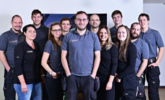 Eine Gruppe von zwölf Personen steht zusammen und lächelt. Sie tragen ähnliche graue Arbeitskleidung und befinden sich in einem hellen Raum. | © Der Elektriker Markus Leitgeb GmbH