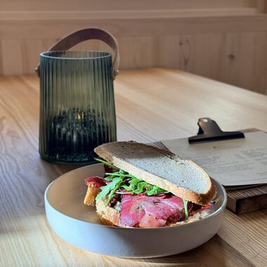 Ein Sandwich mit Rinderbraten und Rucola auf einem Teller. Im Hintergrund steht eine grüne Laterne auf einem Holztisch. | © Einkehr Guntschau