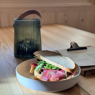 Ein Sandwich mit Rinderbraten und Rucola auf einem Teller. Im Hintergrund steht eine grüne Laterne auf einem Holztisch. | © Einkehr Guntschau