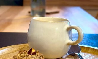 Eine Tasse Kaffee auf einem Holzbrett neben einem Keks. Im Hintergrund ist eine Vase mit bunten Blumen zu sehen. | © Einkehr Guntschau