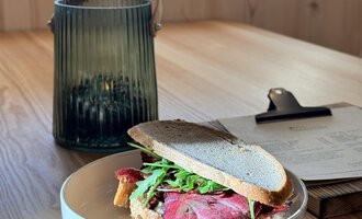 A sandwich with roast beef and arugula on a plate. In the background, there is a green lantern on a wooden table. | © Einkehr Guntschau