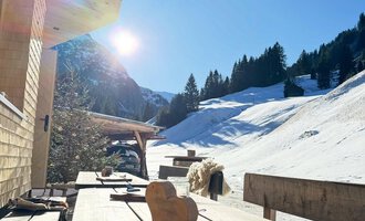 A quiet winter landscape with snow and mountains in the background. On a wooden terrace, some tools are standing, and the sun seems to be shining. | © Einkehr Guntschau