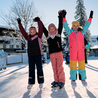 Eine Gruppe fröhlicher Kinder steht auf einer Eislauffläche und hebt die Hände in die Luft. Sie tragen bunte Winterkleidung und genießen den sonnigen Wintertag. | © Kleinwalsertal Tourismus | Oliver Farys