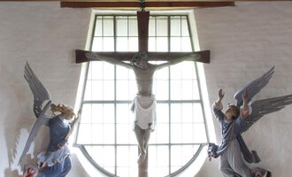 Ein ruhiger Altar in einer Kapelle mit einem Kreuz und Skulpturen von Engeln. Die Fenster lassen viel Licht herein und schaffen eine friedliche Atmosphäre. | © Ev. Kirche Kleinwalsertal