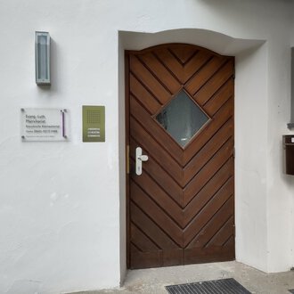 A brown front door with a diamond-shaped window. Next to the door is a mailbox and a sign with information. | © Ev. Kirche Kleinwalsertal