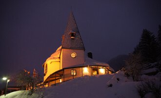 A cozy, snow-covered house with a pointed roof. The lights in the building radiate a warm atmosphere in the wintry night. | © Ev. Kirche Kleinwalsertal