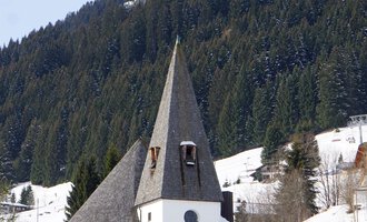 Eine charmante Kirche mit einem spitzen Turm, umgeben von schneebedeckten Wiesen und bewaldeten Bergen. Der Himmel ist klar und die Landschaft wirkt winterlich und friedlich. | © Ev. Kirche Kleinwalsertal