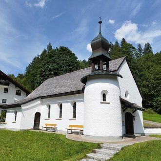 A small, white chapel with a black roof amidst greenery. In the background, trees and a holiday accommodation can be seen. | © Kleinwalsertal Tourismus | N. Lughammer