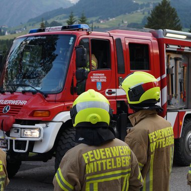 Ein Feuerwehrauto steht auf einer Straße, umgeben von Feuerwehrleuten in uniformen Gelben Helmen. Die Landschaft im Hintergrund zeigt Bäume und Berge. | © Feuerwehr Mittelberg | Christoph Hilbrand
