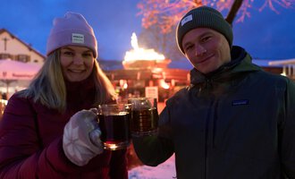 Zwei Personen stoßen mit Gläsern an und lächeln. Im Hintergrund ist eine festliche Atmosphäre mit einer Feuerstelle und 겨우nlichen Lichtern zu sehen.