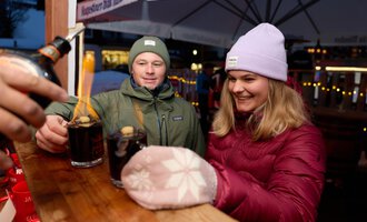 Ein Paar steht an einem Stand und genießt warme Getränke. Beide tragen warme Kleidung und lächeln.