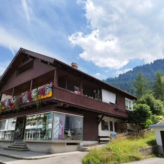 A charming house with a blooming balcony in the mountains. The surroundings are green, and the cloudy sky creates a beautiful atmosphere. | © Kleinwalsertal Tourismus | N. Lughammer
