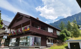 Ein charmantes Haus mit einem blühenden Balkon in den Bergen. Die Umgebung ist grün und die Wolkenhimmel sorgt für eine schöne Atmosphäre. | © Kleinwalsertal Tourismus | N. Lughammer