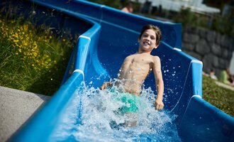 Ein Junge rutscht fröhlich die Wasserrutsche hinunter und spritzt dabei Wasser. Im Hintergrund sind grüne Wiesen und ein paar Schwimmer zu sehen. | © Kleinwalsertal Tourismus | Oliver Farys