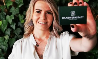 Eine lächelnde Frau hält eine Karte der Haarwerkstatt in der Hand. Im Hintergrund sind grüne Pflanzen zu sehen. | © Friseur Haarwerkstatt | Franziska Diener