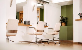 A modern hair salon with elegant white hairdressing chairs and large mirrors. The walls are in neutral colors, with a touch of green accent. | © Friseur Haarwerkstatt | Franziska Diener