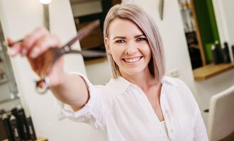 A cheerful hairdresser is holding scissors in her hand and smiling at the camera. Hairdressing elements can be seen in the background. | © Friseur Haarwerkstatt | Franziska Diener