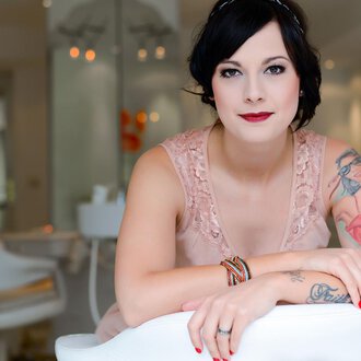 A woman with tattoos is sitting smiling in a modern room. She is wearing an elegant pink dress and has a chic look. | © Fräulein Sugar Coiffeur | Sandra Bromm