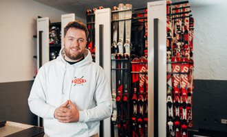 Ein Mann steht lächelnd in einem Skiverleih mit vielen Skiern im Hintergrund. Er trägt einen weißen Hoodie und wirkt einladend und freundlich. | © Friski Marcellus Fritz | Stefan Klauser