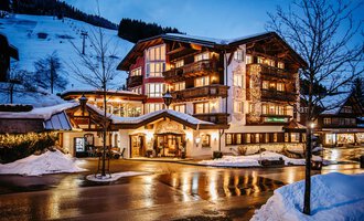 A charming hotel in winter with snow-covered mountains in the background. The warm lights of the house shine in the twilight. | © Hotel Alpenstüble KG