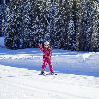 Ein kleines Mädchen in pinker Skiausrüstung fährt fröhlich auf Skiern und winkt. Im Hintergrund sieht man schneebedeckte Bäume und eine verschneite Piste. | © OBERSTDORF · KLEINWALSERTAL BERGBAHNEN