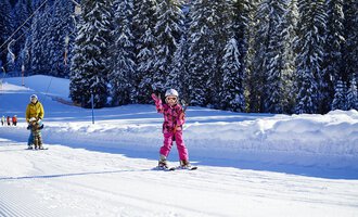 A little girl in pink ski gear is happily skiing and waving. In the background, you can see snow-covered trees and a snowy slope. | © OBERSTDORF · KLEINWALSERTAL BERGBAHNEN