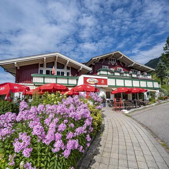 Ein malerisches alpenländisches Haus mit roten Sonnenschirmen und bunten Blumen. Der Himmel ist blau mit einigen Wolken. | © Gasthof Alpenblick | Theodor Pinn